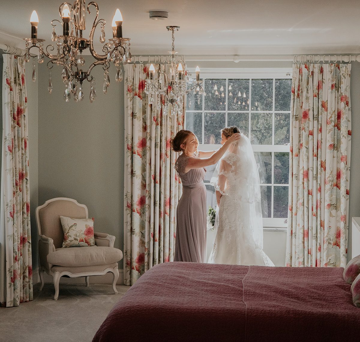 Bride stood by large window in the Bridal Suite at Wooladon Estate alongside her bridesmaid who fixes her veil.