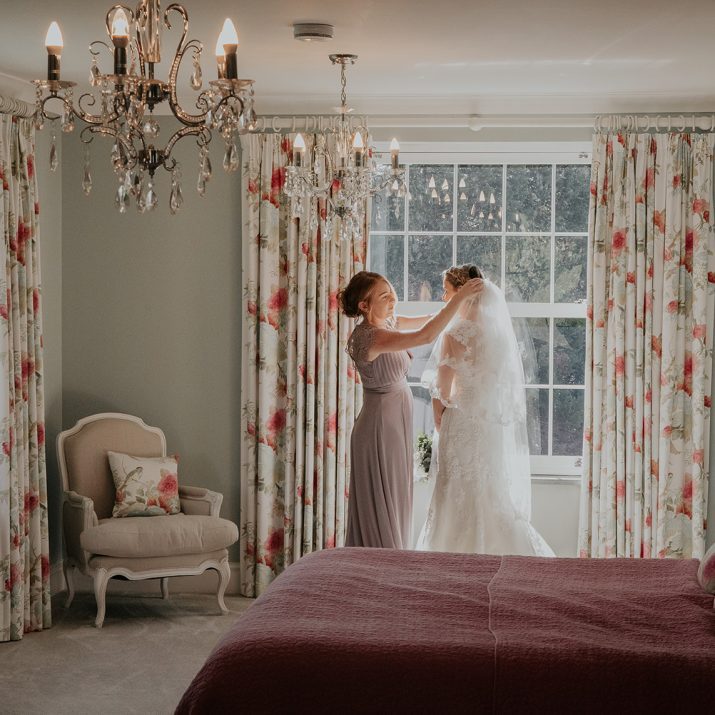 Bride stood by large window in the Bridal Suite at Wooladon Estate alongside her bridesmaid who fixes her veil.